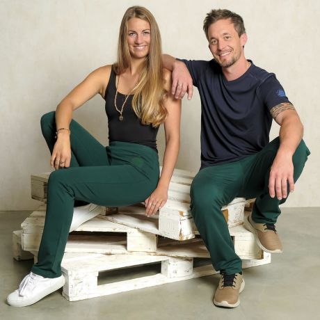 Left woman, right man sitting on white pallets. Bamboo tops and wellness organic cotton pants in fern green. Pants unisex. Flower of life on the hip. Sports pants, yoga pants, sweatpants. Fairly made by The Spirit of OM.