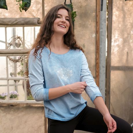 Smiling woman with long hair in 3/4 sleeve shirt Peaceful Lotus from The Spirit of OM. Color sky blue. Blue mandala on the chest, above a silver lotus print. Boho style and effect coloring. Wave embroidery with silver thread on the sleeve. Organic cotton 
