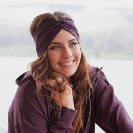 Lächelnde Frau mit offenen, langen Haaren trägt ein Stirnband aubergine oder lila. Material aus Bio-Baumwolle. Kleine Dreiecke sind zu sehen und an der Stirn vorne ist ein Knoten